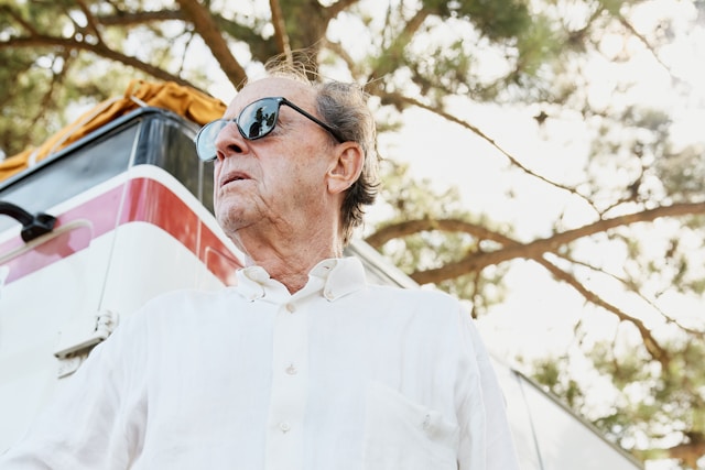 An older man in a white shirt and sunglasses standing beside a vintage camper van under trees